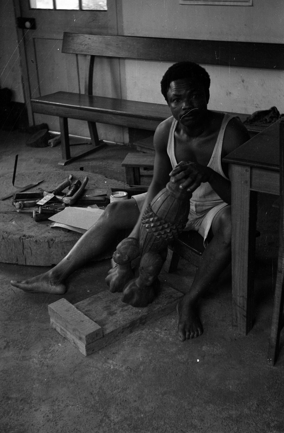 A woodcarver sitting in his workshop shaping a large cylinder of wood, Edo State, 1962
